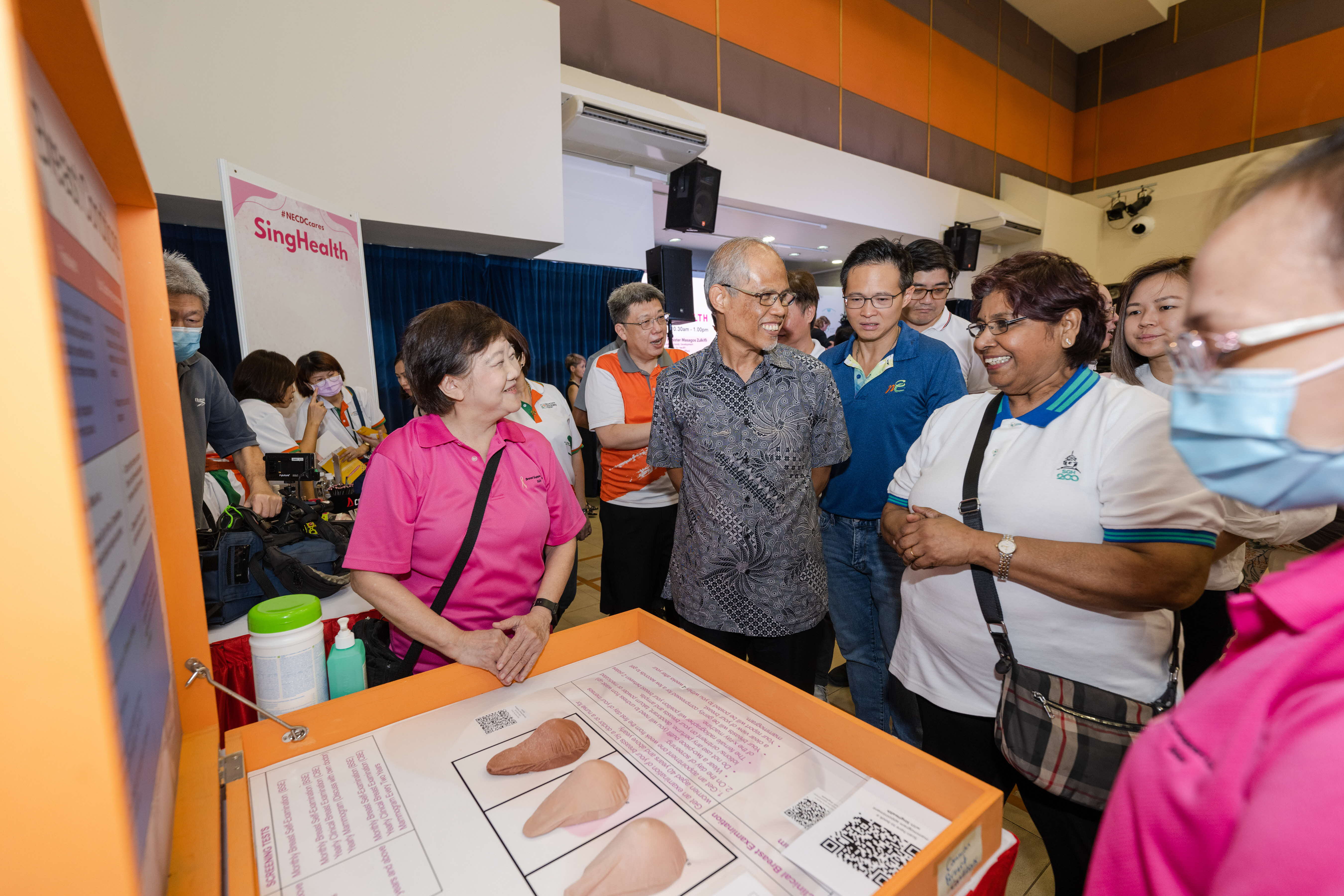 A group of people gather around an informational display booth with SingHealth branding visible in the background.